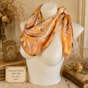 May include: A square silk scarf featuring a vibrant orange and yellow hibiscus floral pattern with subtle striped accents. The scarf includes a white brand tag labeled Oscar de la Renta and a small card stating 100% Silk and 34 x 34 inches.