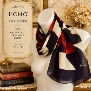 May include: An oblong 100% silk scarf featuring a geometric triangle print in navy blue, red, and cream. The scarf is draped on a white mannequin and displays the brand name ECHO in the corner. The dimensions are listed as 11 inches by 54 inches.