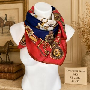 May include: A square silk chiffon scarf featuring a red, navy blue, and cream color palette with a printed equestrian motif of gold chains, buckles, and horse heads. The scarf includes the signature Oscar de la Renta and measures 30 by 30 inches.