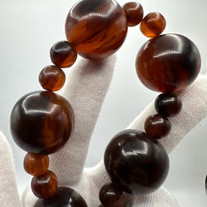 Op de afbeelding: Een close-up van een bruine kralenketting. De ketting heeft grote, ronde kralen in verschillende bruintinten, van licht amber tot donkere chocolade. De kralen zijn aan elkaar geregen, waardoor een opvallend accessoire ontstaat.