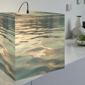 Puede incluir: Una lámpara de mesa cuadrada con un gráfico impreso de agua ondulada en tonos azul, verde y dorado. La lámpara tiene un exterior liso y cúbico, con un cable de alimentación negro, pequeño y delgado, que sale del centro superior del cubo.