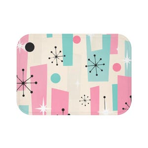 May include: A rectangular bath mat featuring a cream background with a repeating pattern of pink and teal geometric shapes, black starburst icons, white four-pointed stars, and solid circles. The mat has rounded corners and a black circular hole.