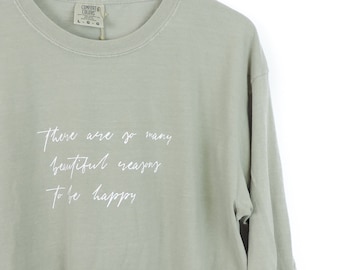 Könnte beinhalten: Salbeigrünes Langarmshirt mit weißem Text, der "Es gibt so viele schöne Gründe, glücklich zu sein" lautet.