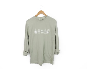 May include: Sage green long-sleeve shirt with a white graphic of various coffee drinks. The shirt is hanging on a wooden hanger against a white background. The sleeves are rolled up.