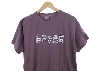 May include: A purple t-shirt with a white graphic of different coffee cups. The graphic includes the word "COFFEE" on one of the cups.