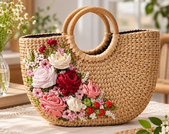 May include: A woven straw handbag with two round handles, featuring an intricate floral arrangement of fabric roses in shades of deep red, light pink, and white, accented with small green leaves and tiny red strawberry embellishments on the front.