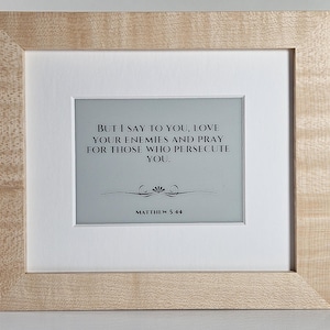 May include: A framed print with a light wood grain frame. The print features the text "BUT I SAY TO YOU, LOVE YOUR ENEMIES AND PRAY FOR THOSE WHO PERSECUTE YOU. MATTHEW 5:44" in black font on a light gray background.