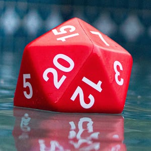 May include: A vibrant red, twenty-sided die with white numerals, partially submerged in water. The die's numbers are clearly visible, with a reflection in the water below. The image captures a gaming accessory.