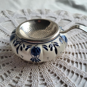 May include: A ceramic tea infuser with a stainless steel strainer and handle. The infuser is white with blue floral designs. The handle is curved. The tea infuser is designed to hold loose tea leaves for steeping.