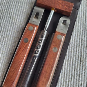 Op de afbeelding: Een bruine leren etui bevat een wijnopener set. De set bevat twee openers met houten handvatten en roestvrijstalen messen, een kurkentrekker en een houten blok. De openers hebben de tekst "ROSTFREI" op de messen.