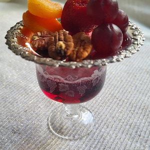 Könnte beinhalten: Ein Stielglas mit tiefroter Flüssigkeit, gekrönt von einer silberumrandeten Schale mit frischen Früchten und Nüssen. Die Anordnung umfasst getrocknete Aprikosen, eine Erdbeere, Trauben und Pekannüsse. Das Glas hat geätzte Blumen- und Traubenmuster.