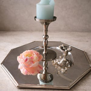 Può includere: Un vassoio ottagonale argentato contiene una candela azzurra in un portacandele decorativo, un fiore rosa e giallo e un piccolo contenitore argentato con una fila di perle. Gli oggetti sono disposti su una superficie grigio chiaro.