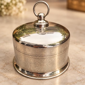 May include: A polished silver-toned circular lidded container with a smooth, reflective finish. The lid features a central vertical handle shaped like a ring attached to a small spherical base, and the body has a simple horizontal line detail.