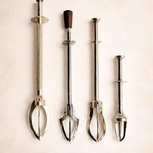 Può includere: Quattro pinze da portata vintage con manici e estremità di presa in metallo. Le pinze variano per dimensioni e design, alcune con un manico strutturato e altre con una parte superiore in legno. Lo sfondo è beige chiaro.