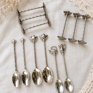 Puede incluir: Una colección de objetos de metal plateado que incluye un estante rectangular con tres barras horizontales, tres soportes verticales con bases circulares y seis cucharas de mango largo. Los mangos de las cucharas tienen remates en forma de tetera.