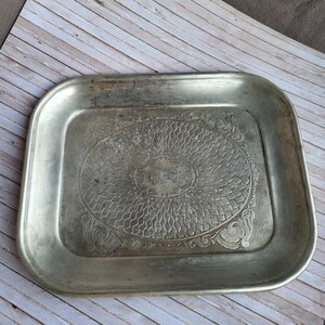 Puede incluir: Una bandeja rectangular de metal plateado con esquinas redondeadas. La bandeja presenta un diseño central ovalado en relieve con un patrón tejido y adornos decorativos en los bordes. La bandeja es probablemente vintage o antigua.