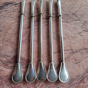 Puede incluir: Cinco bombillas de yerba mate plateadas, cada una con un tallo delgado, un extremo bulboso y perforado, y un anillo decorativo cerca de la parte superior. Las bombillas están dispuestas sobre una superficie marrón texturizada.