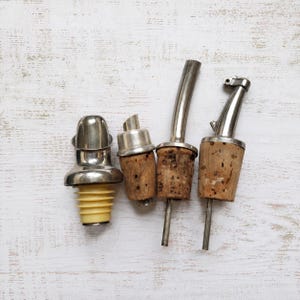Op de afbeelding: Vier flessenstoppers en schenktuiten liggen op een wit houten oppervlak. De stoppers zijn gemaakt van metaal en kurk, met een gele rubberen basis op één. De schenktuiten hebben metalen tuiten en kurken bases.