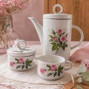 Puede incluir: Un juego de té de porcelana blanca con tetera, azucarero y jarrita para la leche. Cada pieza está decorada con rosas rosas y hojas verdes. La tetera tiene un asa y una tapa con un anillo. El juego está sobre un mantel de encaje blanco.