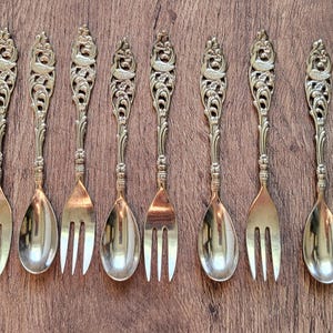 Puede incluir: Un juego de diez utensilios de metal dorado compuesto por cinco cucharas y cinco tenedores. Cada pieza presenta un mango con un diseño intrincado y un motivo de ave, que se conecta a una cabeza de cuchara lisa o de tres dientes.