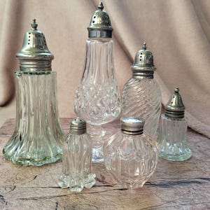 Puede incluir: Una colección de seis saleros y pimenteros de cristal transparente con tapas de metal plateado. Los recipientes presentan diversas formas y texturas, incluyendo estrías verticales, patrones de diamante y diseños en espiral.