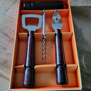 May include: A vintage bar tool set in a box. The set includes a bottle opener, corkscrew, and a bottle cap remover. Each tool has a dark brown wooden handle and a silver metal head. The box is orange and beige.