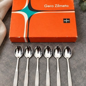 May include: Six silver-toned metal spoons with smooth bowls and vertically ribbed handles, arranged in a row below a rectangular orange box featuring a black and teal star graphic and the white text Gero Zilmeta with a Gero logo.