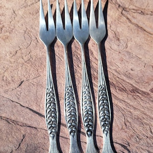 Könnte beinhalten: Ein Set aus vier silberfarbenen Cocktailgabeln aus Metall, jede mit einem zweizinkigen Kopf und einem dekorativen Griff, der mit einem wiederkehrenden Weizenährenmuster geprägt ist und in einer fächerförmigen Basis endet.