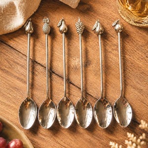 May include: A set of six silver-toned metal spoons with long, slender handles. Each spoon features a unique fruit-shaped finial at the top, including designs of a pear, pineapple, and other assorted fruits, with polished, reflective bowls.