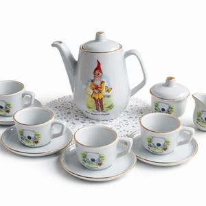 May include: A white ceramic tea set with gold trim, featuring a teapot with a gnome illustration, six teacups and saucers, a sugar bowl, and a creamer. The set is arranged on a white doily.