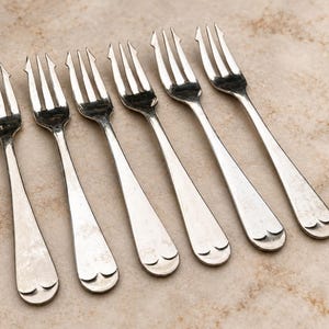 Könnte beinhalten: Ein Set aus sechs silberfarbenen Metallgabeln, jede mit drei Zinken und einem Griffende, das mit einem kleinen, geprägten herzförmigen Detail verziert ist. Die Gabeln sind in einer parallelen Reihe angeordnet und haben eine polierte Oberfläche.