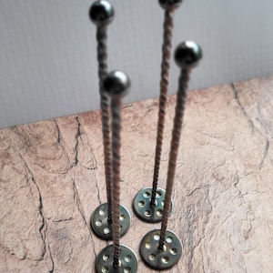 May include: Four metal insulation fasteners. Each has a round base and a twisted rod, topped with a small, dark sphere. They are on a textured, brown surface.
