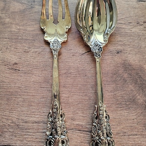 Pode incluir: Um conjunto de garfo e colher dourados para servir. O garfo tem quatro dentes e a colher tem uma tigela grande e oval. Ambos os utensílios apresentam cabos ornamentados e detalhados com desenhos florais e de rolagem, perfeitos para servir em um jantar formal.