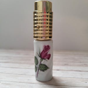 Peut inclure: Un flacon de parfum décoratif avec un dessus doré et une base blanche. La base présente un motif peint d'une rose rouge avec des feuilles vertes. Le dessus est cylindrique avec un design nervuré.