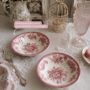 Può includere: Due ciotole ovali in ceramica con un motivo floreale rosso e bianco. Ogni ciotola misura 15 cm di diametro e 4 cm di altezza. Le ciotole sono su una tovaglia di pizzo bianco, con una forchetta vintage e un tovagliolo rosa.