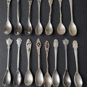 May include: A collection of fifteen silver-toned metal spoons arranged in two rows. Each spoon features a unique decorative handle, with several displaying embossed letters including A, B, C, D, G, J, I, K, L, and M, alongside intricate filigree patterns.