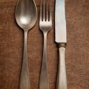 Peut inclure: Gros plan sur une cuillère, une fourchette et un couteau en argent, disposés sur une surface en bois. Les couverts présentent une finition argentée et un design classique. La surface en bois est de couleur marron chaud.