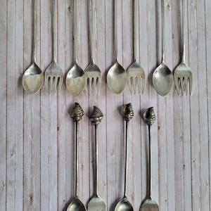 Può includere: Una collezione di cucchiai e forchette argentati con manici decorativi. I manici presentano vari disegni a conchiglia e a spirale. Le posate sono disposte su una superficie di legno chiaro, mettendo in risalto i loro dettagli intricati e lo stile elegante.