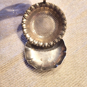 Könnte beinhalten: Ein Teesieb aus silberfarbenem Metall mit einer sonnenblumenförmigen Schale, die ein perforiertes Zentrum aufweist, sowie eine passende Tropfschale mit gewelltem Rand. Das Sieb hat einen einzelnen vertikalen Griff am oberen Rand.