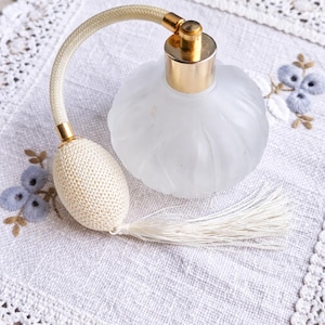 Pode incluir: Um frasco de perfume vintage de vidro fosco com uma tampa dourada e um borrifador de bulbo creme. A garrafa tem formato arredondado com ranhuras verticais. O borrifador tem um bulbo e borla tecidos. A garrafa repousa sobre um tecido branco bordado.