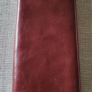 May include: A burgundy leather wallet, rectangular in shape, with a smooth, slightly glossy finish. The wallet is closed and appears to be empty. The edges are neatly stitched. The wallet is lying on a textured surface.