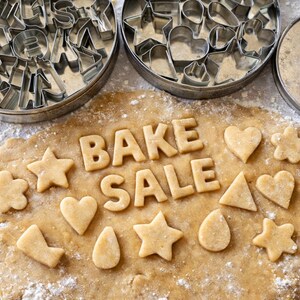 Könnte beinhalten: Eine Backszene mit Ausstechern und Teig. Die Worte "BAKE SALE" sind in Buchstaben aus Keksen geschrieben. Verschieden geformte Kekse, darunter Sterne, Herzen und Blumen, sind um den Text herum verstreut. Zwei Metalldosen mit Ausstechern befinden sich im Hintergrund.