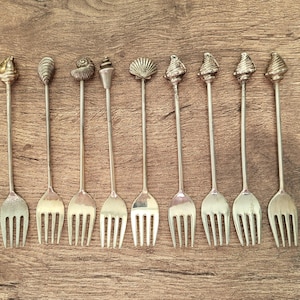 Può includere: Un set di nove forchette da antipasto in metallo color argento, ognuna caratterizzata da un terminale unico a forma di conchiglia o ispirato al mare sulla parte superiore del manico. Le forchette hanno steli lunghi e sottili e quattro rebbi.