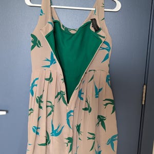 Pu&ograve; includere: Un abito senza maniche con fodera verde ed esterno beige. L'abito ha una chiusura a zip e presenta un motivo di uccelli blu e verdi.