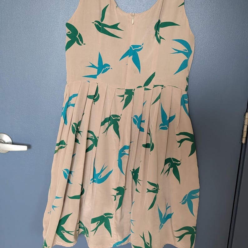 Pu&ograve; includere: Un abito beige senza maniche con stampa di uccelli blu e verdi. L'abito ha una gonna plissettata e un corpetto aderente.