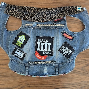 May include: A blue denim dog vest with patches, including "Black Dog", "Kennel Your Idols", "Drop Treats Not Bombs", "Foodie", and "Non Human Animalz". The vest has a leopard print collar and silver studs.