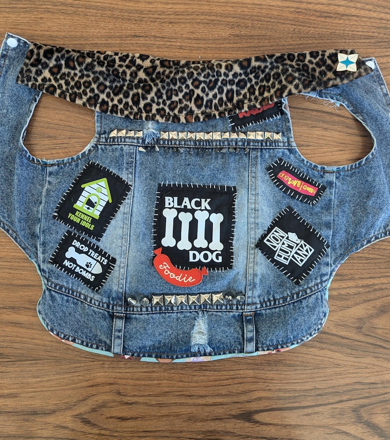 May include: A blue denim dog vest with patches, including "Black Dog", "Kennel Your Idols", "Drop Treats Not Bombs", "Foodie", and "Non Human Animalz". The vest has a leopard print collar and silver studs.