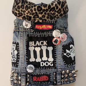 May include: A blue denim vest with a leopard print collar, studded with silver spikes. The vest is decorated with patches and pins, including the text "BLACK DOG" and "RABID".
