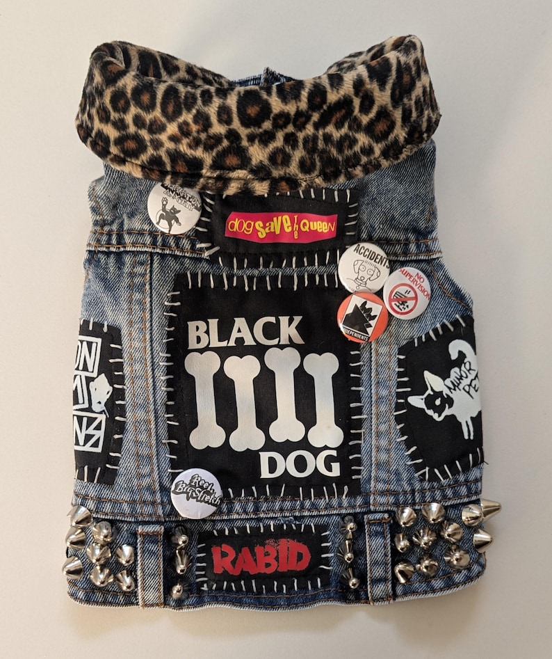May include: A blue denim vest with a leopard print collar, studded with silver spikes. The vest is decorated with patches and pins, including the text "BLACK DOG" and "RABID".