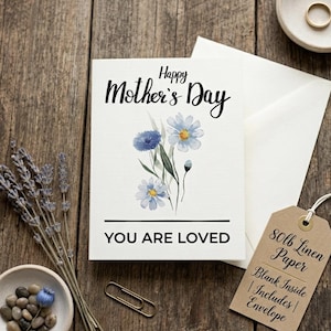 May include: A Mother's Day card with blue watercolor flowers and the text "Happy Mother's Day" and "You Are Loved." The card is on a wooden surface with a white envelope, a tag, and lavender sprigs.